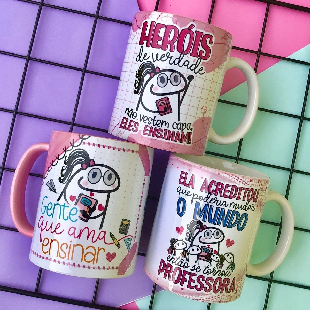Tres canecas estampadas com ilustracoes divertidas e frases sobre professores dispostas sobre grade colorida.