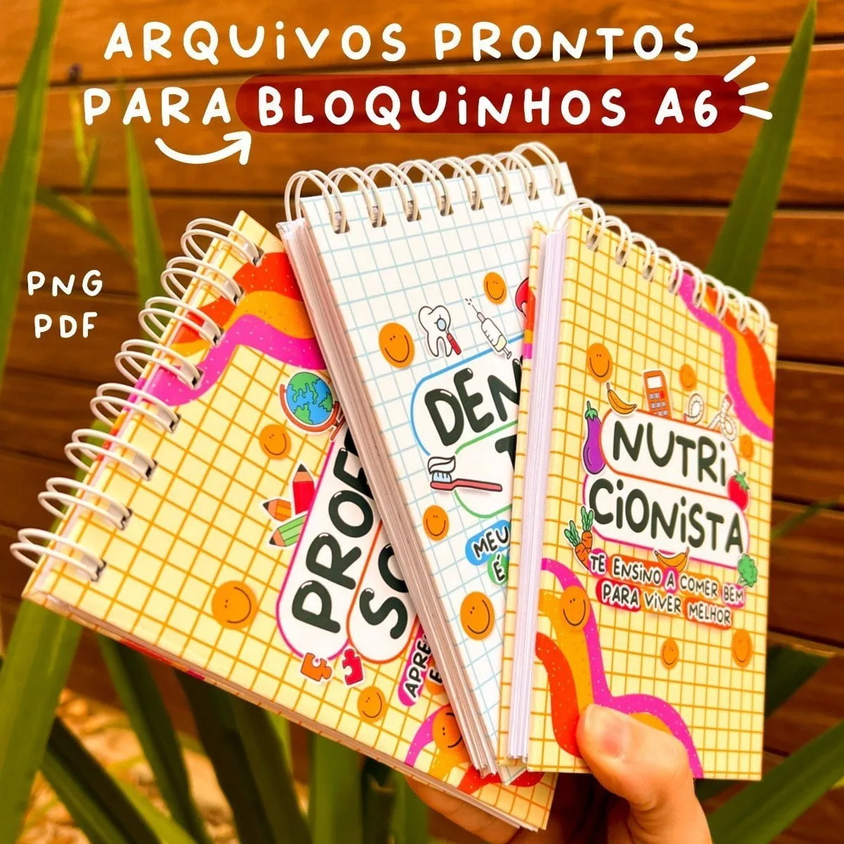 Três cadernos espiralados com capas coloridas decoradas sobre fundo de madeira e folhagens.