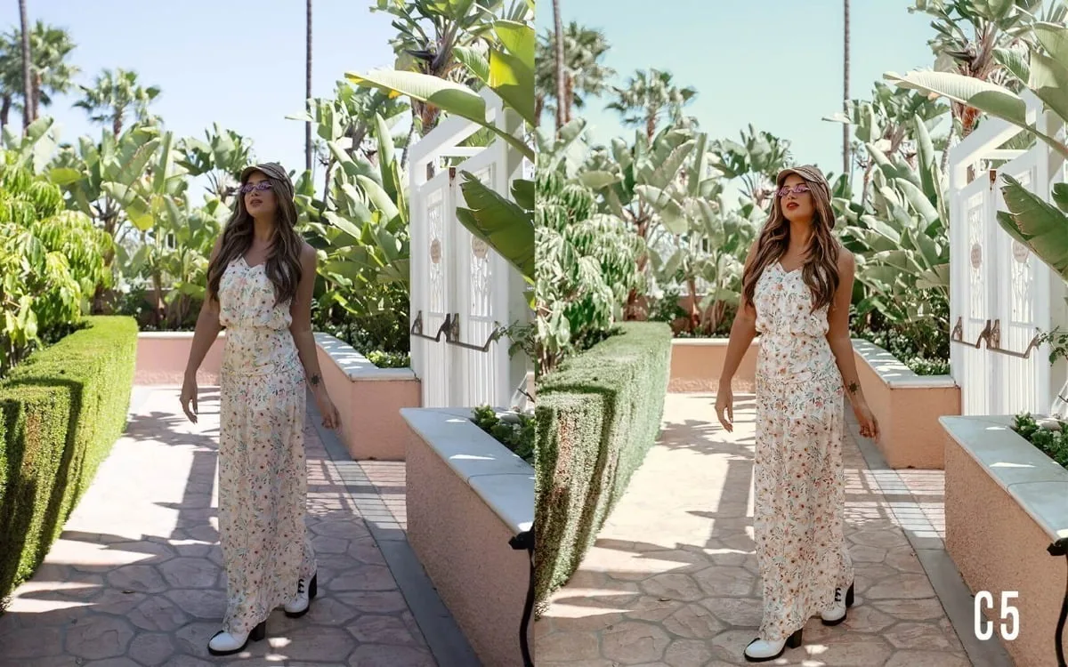 Mulher de vestido floral e chapéu caminha em jardim ensolarado com palmeiras ao fundo.