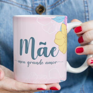 Caneca rosa com "Mãe meu grande amor" e flores coloridas, segurada por mãos.