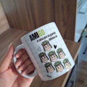 Mão segurando uma caneca branca com ilustrações de personagem e texto em português.