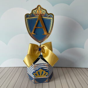 Peça decorativa de festa com escudo azul e dourado, letra A, laço e coroa.