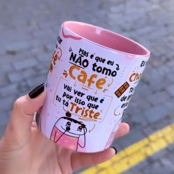 Mão com unhas pretas segura caneca branca e rosa estampada com texto e desenho.