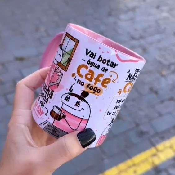 Mão segura uma caneca branca e rosa estampada com desenhos e textos divertidos.