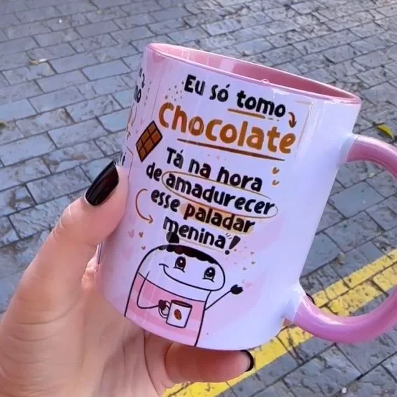 Mão segurando caneca branca e rosa com estampa de frases e desenho.