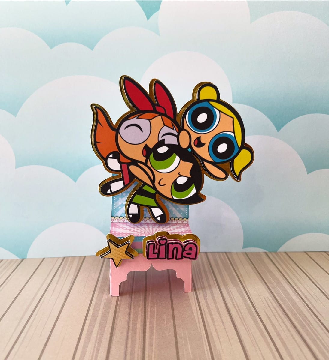 Kit Festa Meninas Super Poderosas em Studio e PDF 2 Três personagens animados coloridos, um com laço vermelho, em um suporte decorado.