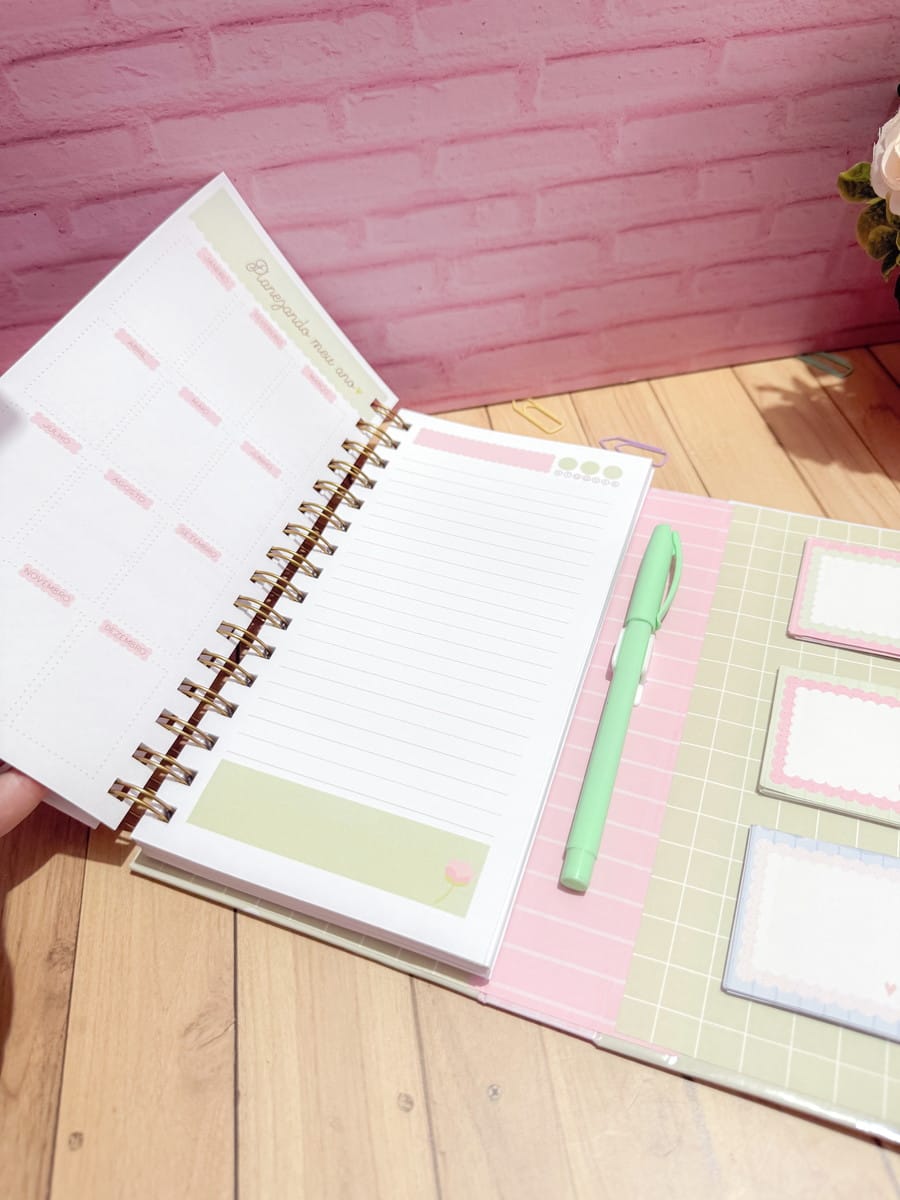 Kit Agenda Floral 2026 para Imprimir e Encadernar 1 Caderno aberto com espiral dourado, caneta verde e blocos adesivos coloridos sobre madeira.