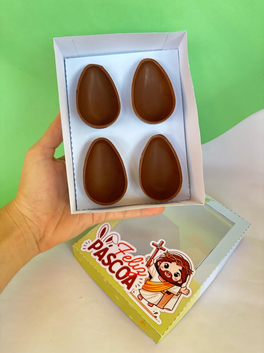 Kit Caixas Páscoa Canva para Corte em Plotter 4 Mão segura caixa branca com quatro metades de ovos de chocolate. Adesivo "Feliz Páscoa" visível.