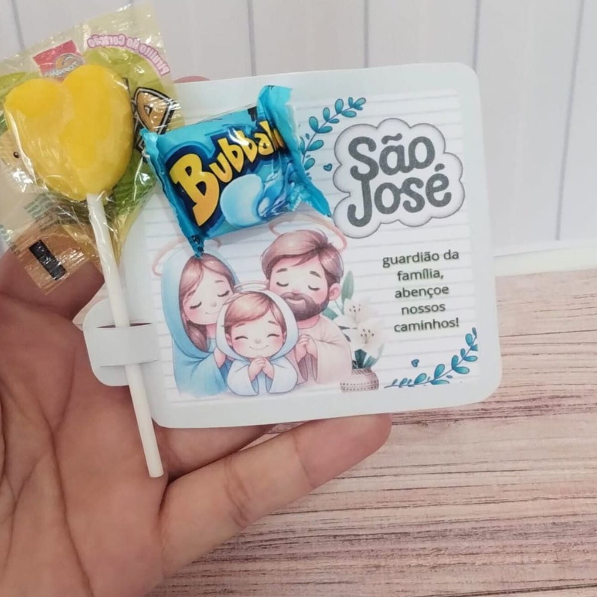Kit Digital Lembrancinha São José para Imprimir e Cortar 4 Mão segurando cartão impresso com ilustração religiosa, pirulito amarelo e bala azul.