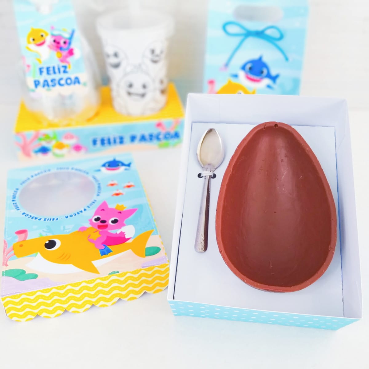 Kit digital Páscoa Baby Shark com caixas e sacolinhas 2 Composição visual de Páscoa com meio ovo de chocolate, colher e embalagens temáticas.