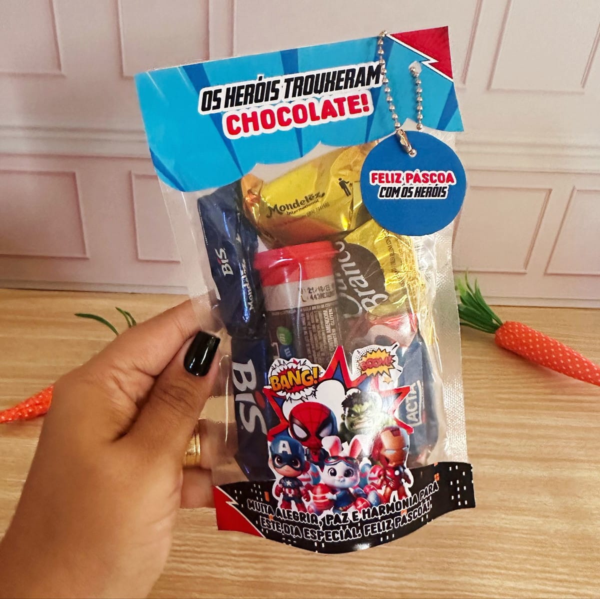 Kit Digital Páscoa Super Heróis para Corte e Impressão 5 Sacola transparente de Páscoa com chocolates variados, tema de heróis, segurada por mão.