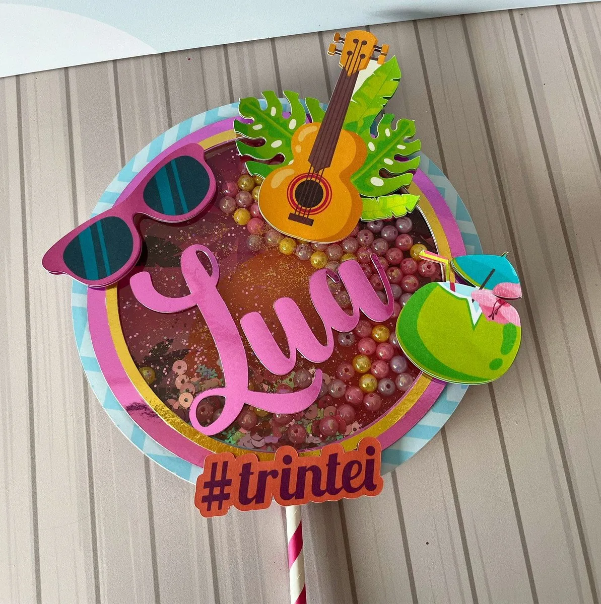 Topo de bolo circular decorado com tema tropical, violão, óculos e coqueiro pequeno.