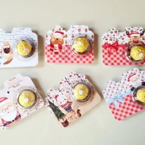 Seis cartões decorados com motivos natalinos contendo bombons redondos embrulhados em papel dourado.